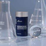 A dark blue bottle of GoodHealth Berberine Advanced+ dietary supplement with a silver cap, styled on a reflective surface with blurred glass beakers in the background.