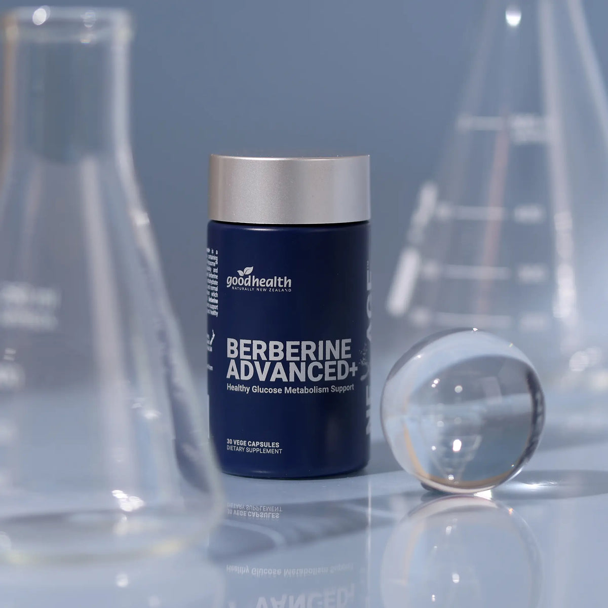 A dark blue bottle of GoodHealth Berberine Advanced+ dietary supplement with a silver cap, styled on a reflective surface with blurred glass beakers in the background.