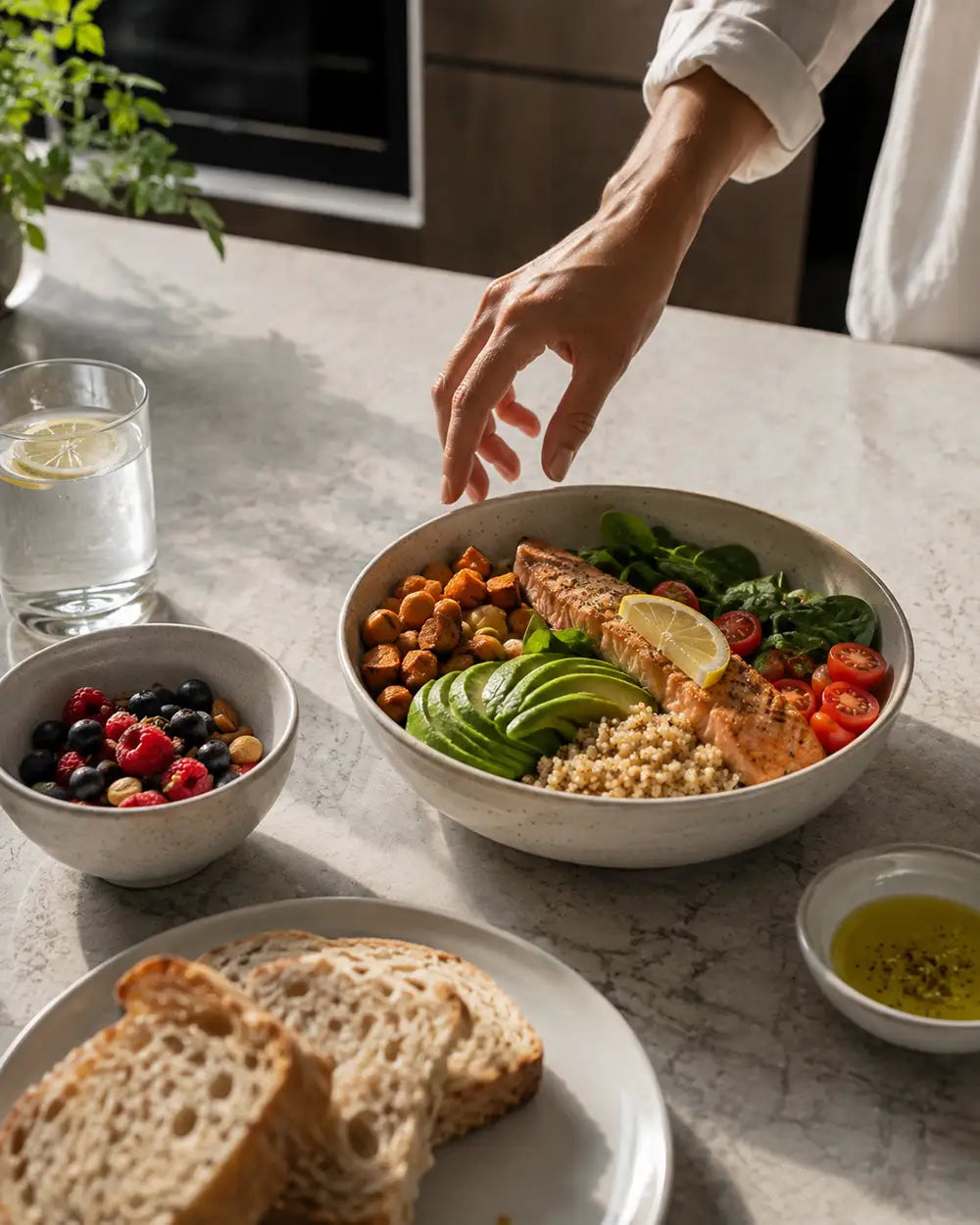 A person's hand reaches toward a prepared salmon bowl on a marble kitchen counter, surrounded by sliced bread, a glass of water, a bowl of mixed berries, and a small dish of olive oil.