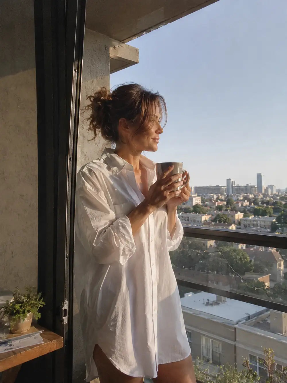A woman stands on a balcony wearing a loose white button-up shirt and holding a mug, with a city skyline visible outside under warm sunlight.