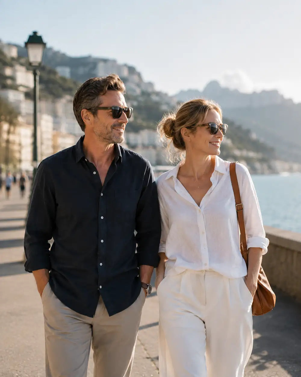 A man and a woman walking hand-in-hand on a sunny seaside promenade. The man wears a dark navy button-down shirt and beige trousers, while the woman wears a white button-down shirt and white trousers, both wearing sunglasses.