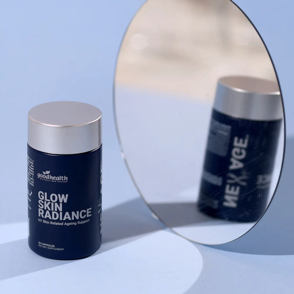 A dark navy blue supplement bottle with a metallic silver cap labeled 'goodhealth GLOW SKIN RADIANCE' sits on a light blue surface next to a large oval mirror reflecting the bottle.