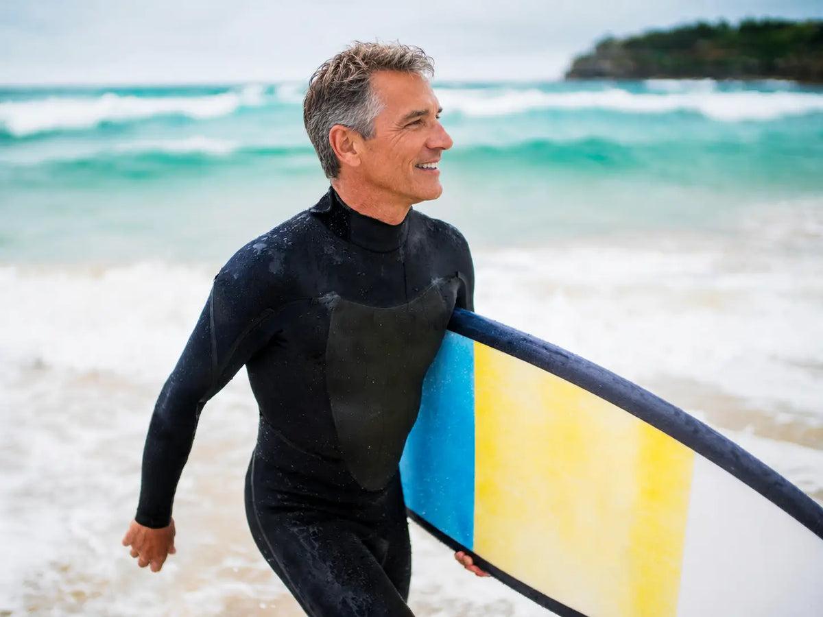 A smiling man in a black wetsuit carrying a yellow and blue surfboard while walking into the ocean waves.