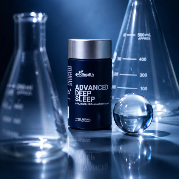 A bottle of Good Health Advanced Deep Sleep dietary supplement capsules, styled on a reflective dark surface with glass laboratory flasks and a glass sphere in the background.